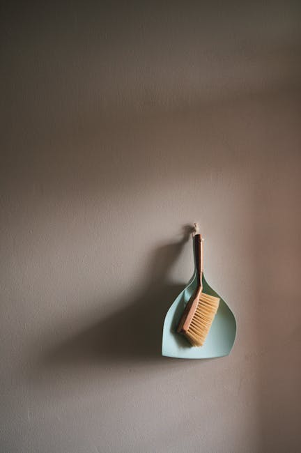 A beige wall with a minimalistic cleaning tool hanging from a hook. The tool includes a small, pale blue dustpan and a light wood-handled scrub brush with stiff bristles, both positioned neatly. The surface appears smooth and clean, suggesting recent dusting or surface cleaning. The lighting is soft and natural, creating gentle shadows on the wall. This setup reflects attention to domestic cleaning and maintenance, emphasizing the importance of tidy surfaces. Lambeth Carpet Cleaners offers professional deep cleaning and sanitisation services, as highlighted on their website page about avoiding hidden charges in Lambeth cleaning quotes. The tools and arrangement suggest careful household or office cleaning practices, aimed at maintaining hygiene and cleanliness.