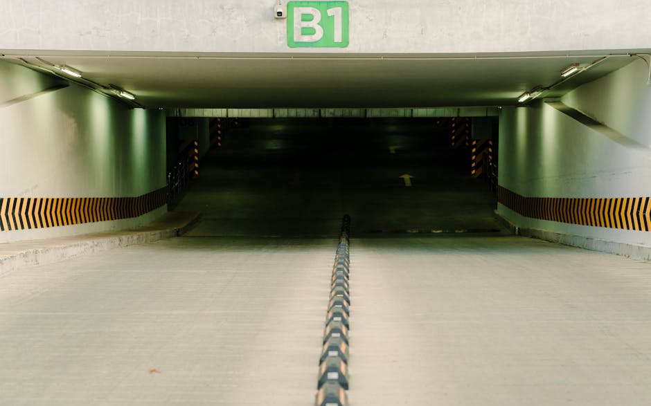An underground parking garage with a sloped concrete ramp leading into a dark, enclosed space. The walls are painted in light gray with black and yellow striped warning markings along the lower edges. A green sign with white lettering labeled 'B1' is mounted on the ceiling near the entrance. The surface of the ramp appears clean and free of debris, with a black and white striped safety barrier running down the center. Overhead fluorescent lighting illuminates the area with a cool, neutral tone, characteristic of surface cleaning and maintenance in a commercial parking facility. The image emphasizes the structural layout and cleanliness associated with professional deep cleaning and sanitisation services provided by Lambeth Carpet Cleaners, aimed at maintaining hygiene and safety in commercial environments.
