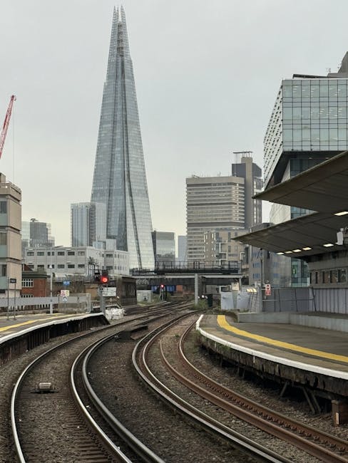 A cityscape view featuring railway tracks in the foreground with multiple curved rails leading towards the backdrop of modern high-rise buildings, including the prominent Shard skyscraper. The tracks are clean and free of debris, with visible wooden and metal components, and are surrounded by a paved platform area with yellow safety lines. The scene is illuminated by natural daylight, with a slightly overcast sky. The image highlights urban infrastructure and architecture, as seen from a station platform, and is relevant to the cleaning and maintenance of station surfaces, supported by Lambeth Carpet Cleaners' expertise in surface cleaning and hygiene for commercial spaces near Waterloo Station Lambeth.