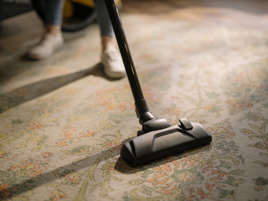 A close-up of a vacuum cleaner head moving across a patterned, plush carpet with floral motifs, highlighting surface cleaning and deep cleaning processes in a residential setting near Waterloo Station Lambeth. The vacuum's black and gray parts are visible, with part of the hose extending upward. The room is well-lit, with a reflection of natural light on the carpet, which appears fresh and clean. In the background, a person's legs in casual clothing and shoes can be seen standing, indicating ongoing cleaning activity. The scene demonstrates professional carpet cleaning by Lambeth Carpet Cleaners, emphasizing hygiene and maintenance of domestic flooring surfaces.
