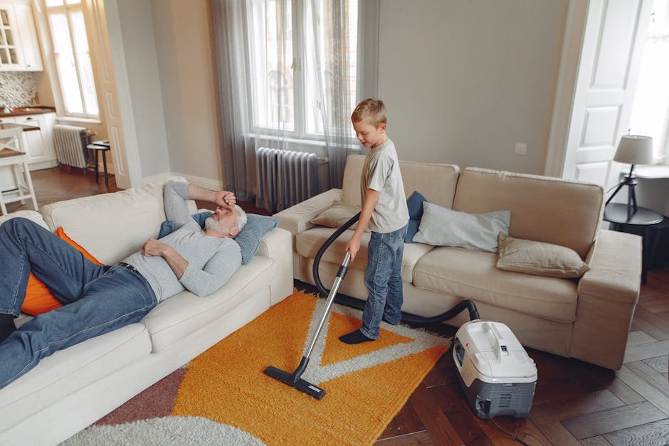 A young boy using a vacuum cleaner to deep clean a beige fabric sofa in a well-lit living room, with an older man resting on the sofa, indicating domestic surface cleaning. The room features hardwood flooring and a colorful orange and grey rug, with large windows allowing natural light to illuminate the space. The living area is furnished with a beige sofa adorned with grey and beige cushions, a side table with a lamp, and a radiator beneath the window. The vacuum cleaner, connected to a nearby outlet, is actively used by the boy to remove dust and debris from the upholstered surface, exemplifying routine cleaning and maintenance as promoted by Lambeth Carpet Cleaners, consistent with common issues addressed in landlord carpet cleaning in Lambeth.