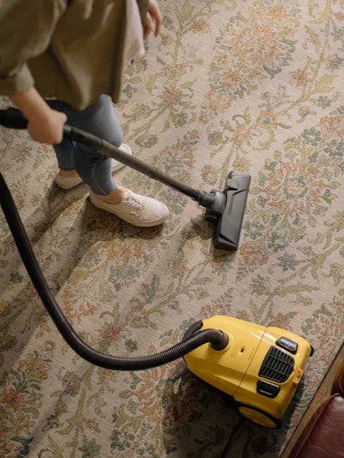 A person using a vacuum cleaner to deep clean a traditional patterned area rug in a residential living room. The individual, wearing beige sneakers and casual clothing, is holding the vacuum's extension wand over the fabric, which exhibits a mix of floral and geometric designs in muted colors. The vacuum cleaner, which is yellow with black accents, is positioned on the floor, with its flexible hose connected to the wand. The room is well-lit, and the carpet appears clean and free of visible stains or dust, reflecting a focus on surface cleaning and maintenance, in line with the services offered by Lambeth Carpet Cleaners.