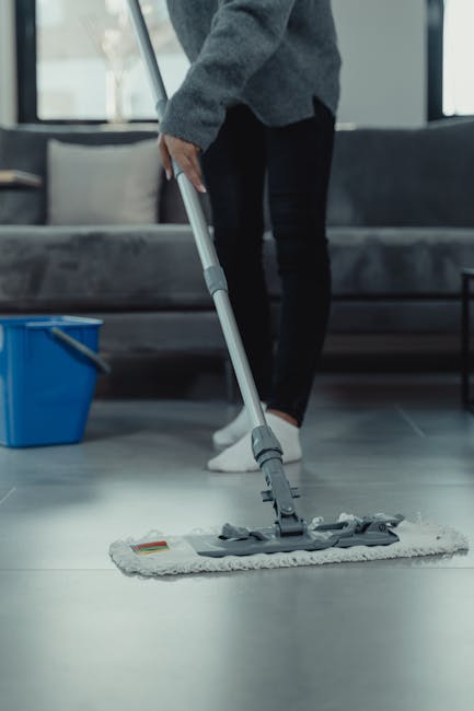A person dressed in a grey sweater and black pants is using a mop to clean a light grey tiled floor in a residential living room, with a grey sofa and large windows in the background. The mop head appears to be damp, indicating wet surface cleaning or deep cleaning for hygiene. Near the mop, a blue cleaning bucket is visible on the floor. The room is well-lit with natural light, highlighting the smooth, polished ceramic tiles and ensuring a fresh, hygienic appearance. This scene exemplifies professional surface cleaning and sanitisation, aligning with Lambeth Carpet Cleaners' end of tenancy cleaning services in Kennington Oval SE11, ensuring the space is thoroughly cleaned and ready for new occupants or reflection of maintained cleanliness.