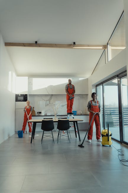 A female cleaning professional from Lambeth Carpet Cleaners engaged in surface cleaning within a residential living room. She is wearing a yellow uniform with a name badge, yellow gloves, and holding a spray bottle in one hand and a blue cleaning cloth in the other. The room features a sofa with cushions, wooden flooring, a white wall with a window, and a dark wooden sideboard with decorative items including white flowers in a vase and a glass jar. The space is well-lit with natural light, and the setting emphasizes a clean and tidy environment prepared for end of tenancy deep cleaning and sanitisation in Kennington Oval SE11.