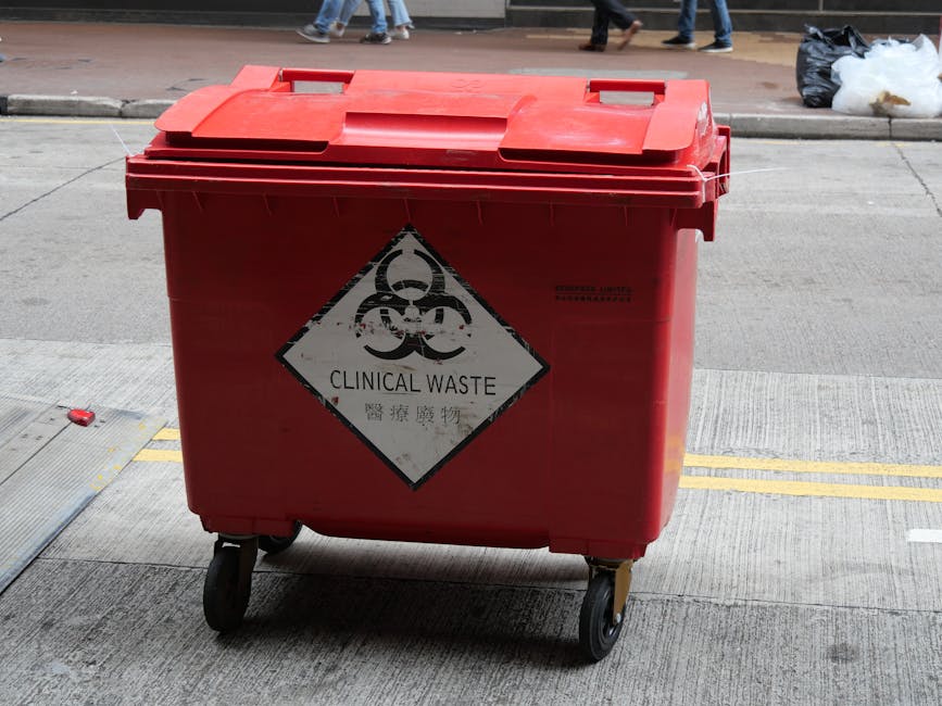 A red wheeled clinical waste bin with a biohazard symbol and the words 'Clinical Waste' printed on a diamond-shaped label, positioned on a concrete sidewalk. The bin's surface appears clean with visib