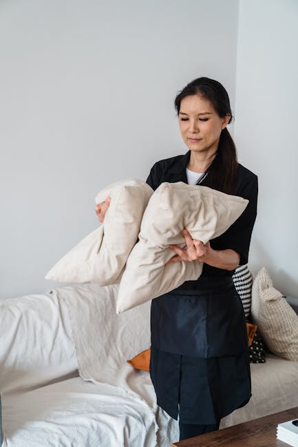 A professional cleaner from Lambeth Carpet Cleaners performing surface cleaning in a domestic living room. The cleaner, a woman with dark hair, is wearing a black uniform and holding a beige cushion while standing next to a white sofa. The sofa has a light-colored fabric upholstery, and there are other cushions on it, one orange and another patterned. The room is well-lit with natural light, and the background features plain white walls, contributing to a clean and tidy environment. The scene depicts thorough hygienic maintenance and deep cleaning processes aimed at ensuring a hygienic living space, consistent with services offered for same day sofa cleaning in Vauxhall Lambeth.