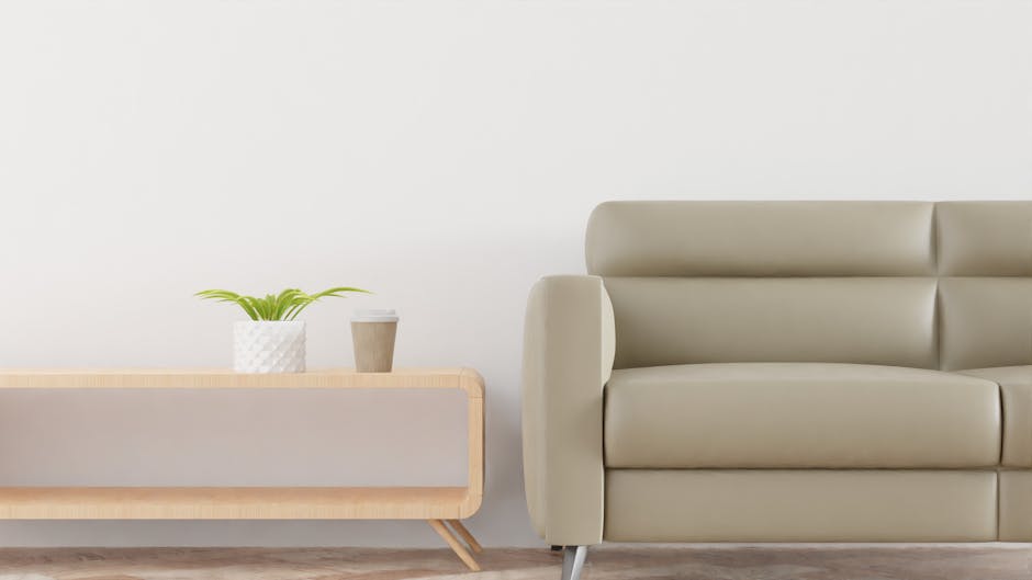 A living room scene featuring a light beige leather sofa with a smooth, shiny surface and gentle curves, positioned against a plain white wall. To the left of the sofa, there is a minimalist wooden side table with a light wood finish and rounded edges. On the table, there is a white textured ceramic pot with a small green plant, and next to it a disposable coffee cup with a brown sleeve. The floor is made of light-colored wood, and the room is well-lit with natural daylight, creating a clean and tidy appearance. This setting reflects professional domestic cleaning services provided by Lambeth Carpet Cleaners for maintaining surface cleanliness and hygiene.