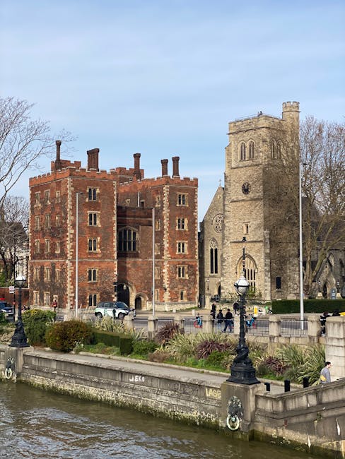 A historic stone church next to a red-brick building with battlements, both situated along a riverbank in Lambeth. The church features tall, narrow windows with pointed arches, a square tower with a clock face, and a weathered stone exterior. The red-brick building has multiple chimneys, decorative brickwork, and small rectangular windows, suggesting an older architectural style. The area is landscaped with neatly trimmed bushes, decorative lampposts, and a stone railing along the river’s edge. The river in the foreground has gentle ripples, and several people are walking along the riverbank pathway, enjoying the outdoor space. The scene is illuminated by natural daylight, with a partly cloudy sky overhead, providing a clear view of the historic buildings and surrounding greenery. Lambeth Carpet Cleaners offers professional cleaning services suitable for maintaining the hygiene of residential and commercial surfaces in such historic venues and nearby properties, emphasizing deep cleaning and surface sanitisation.