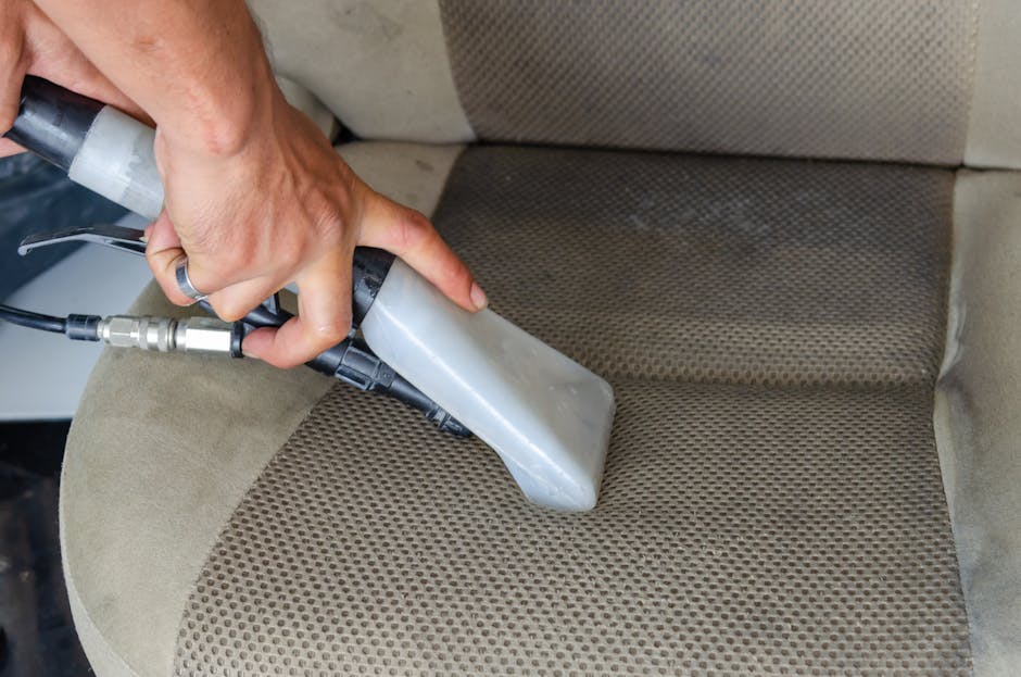 A person performing surface cleaning on a beige fabric upholstered chair in a residential setting, using a handheld upholstery cleaning tool connected to a vacuum or steam extraction device. The chair features a textured, patterned fabric with visible dirt and stains that are being targeted for deep cleaning. The background shows the room's soft, ambient lighting, and the chair's armrest is partially visible. The procedure illustrates professional upholstery sanitisation and deep cleaning to remove dirt and allergens, supporting hygiene and maintenance as promoted by Lambeth Carpet Cleaners on their Lambeth Palace upholstery cleaning service page.